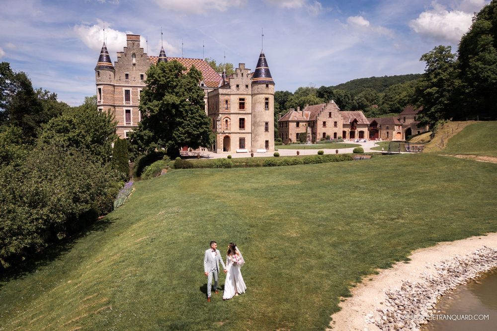 Couple in wedding attire walking on green lawn before a grand castle surrounded by lush trees on a sunny day.