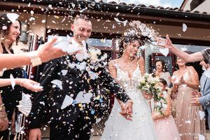 Smiling bride and groom hold hands as guests toss confetti during their outdoor wedding celebration.