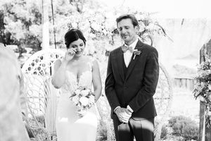 Black and white photo of an emotional bride wiping a tear beside a smiling groom during an outdoor wedding ceremony with floral decor.