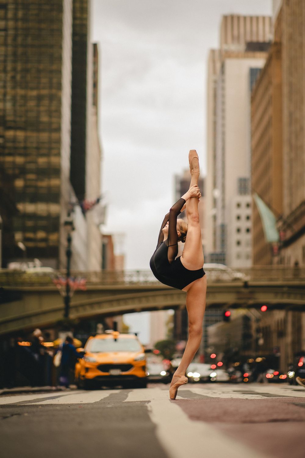 A dancer performs a graceful leap across a New York City street with yellow taxis and skyscrapers in the background.