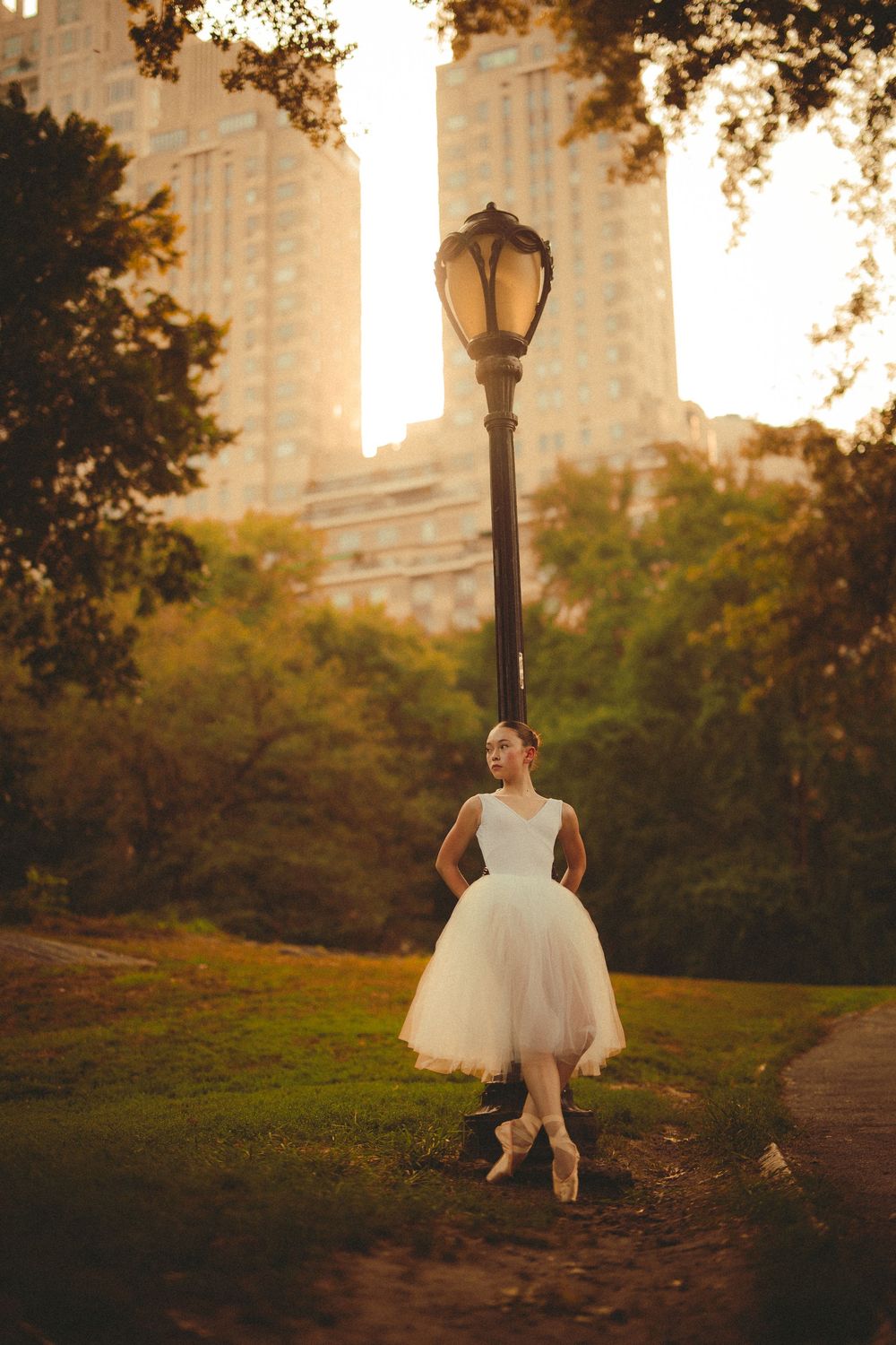 Ballet dancer in white tulle dress poses en pointe by vintage lamppost in Central Park at golden hour.