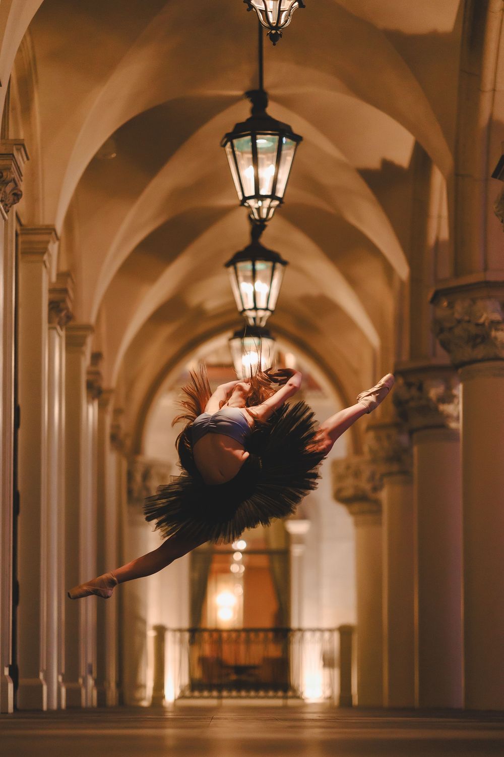 Ballet dancer leaping gracefully through an elegant arched hallway illuminated by hanging lanterns.
