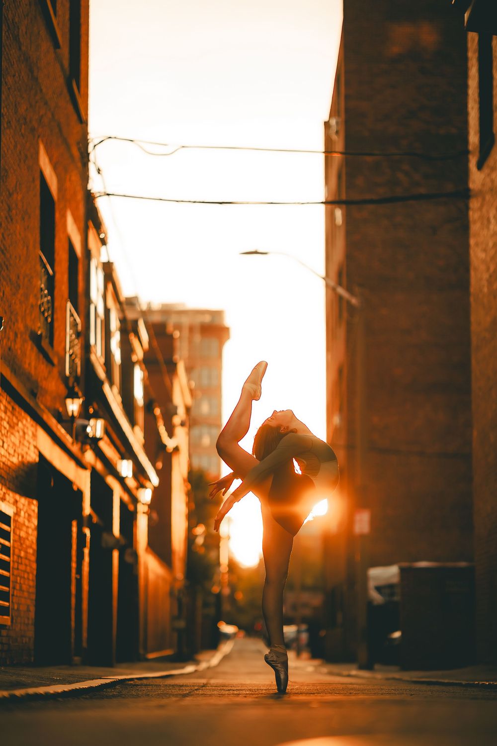 A ballet dancer performs an elegant pose on a city street at sunset, silhouetted against the warm golden light.