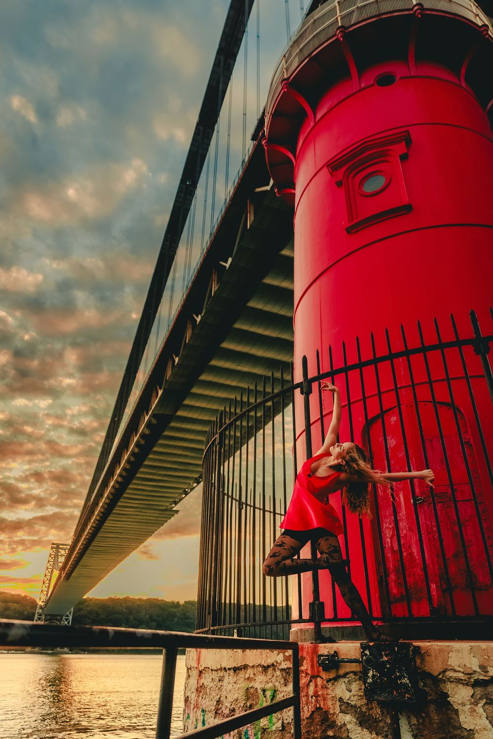 A red lighthouse against a dramatic sunset sky with clouds and metal railings in the foreground.