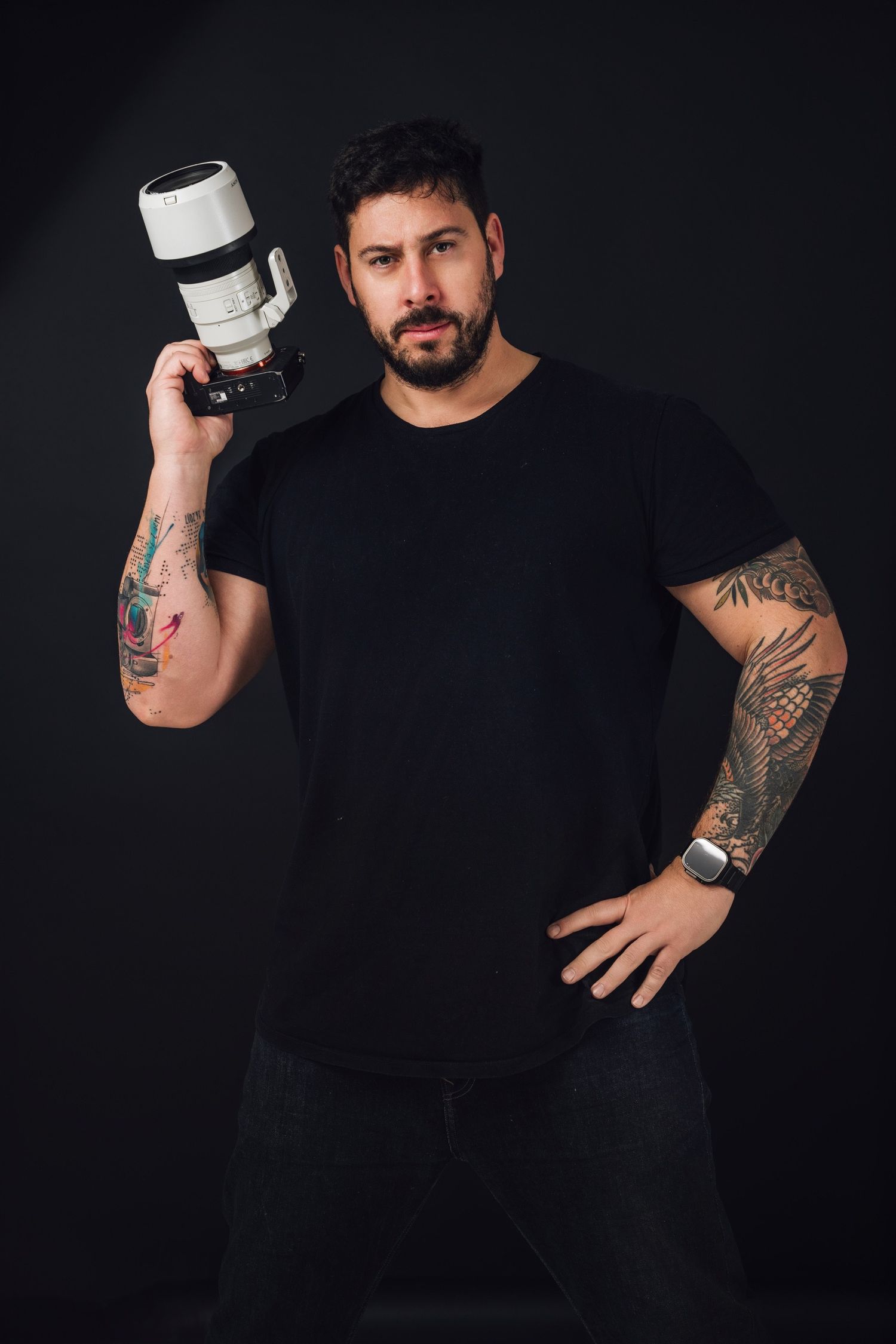 Tattooed photographer in black shirt posing with camera lens against dark background.