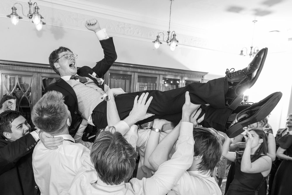Groom being lifted by wedding guests during a lively reception celebration in a grand ballroom, black and white photo.