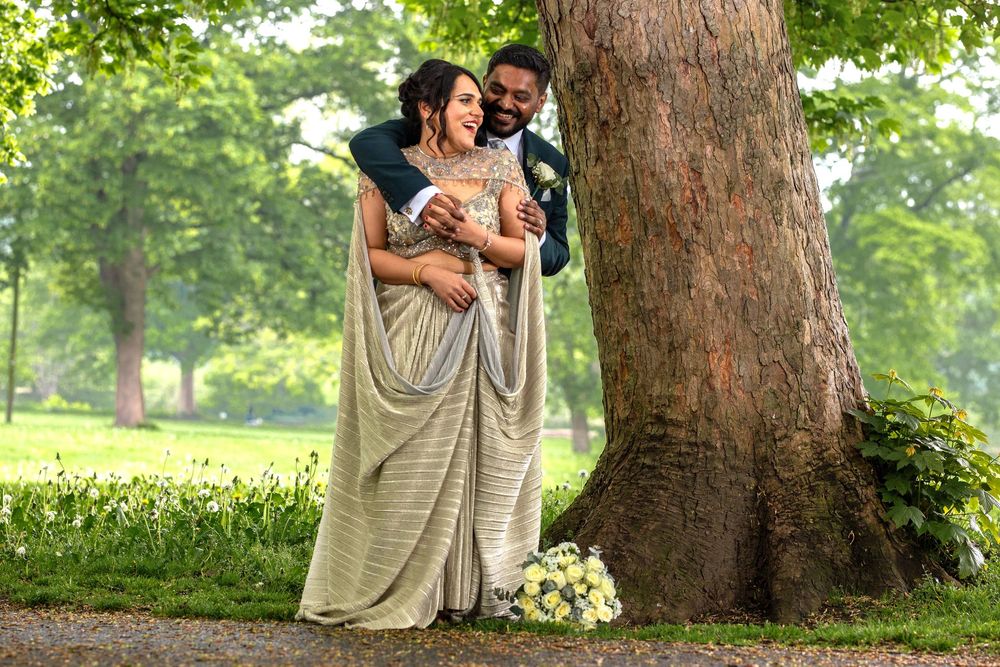 Bride in gold sari and groom in suit embrace by a tree in a lush green park on their wedding day.