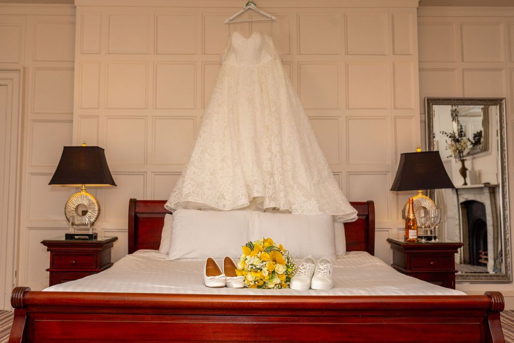 A white wedding dress hangs above a bed with yellow flowers and gold shoes arranged below.