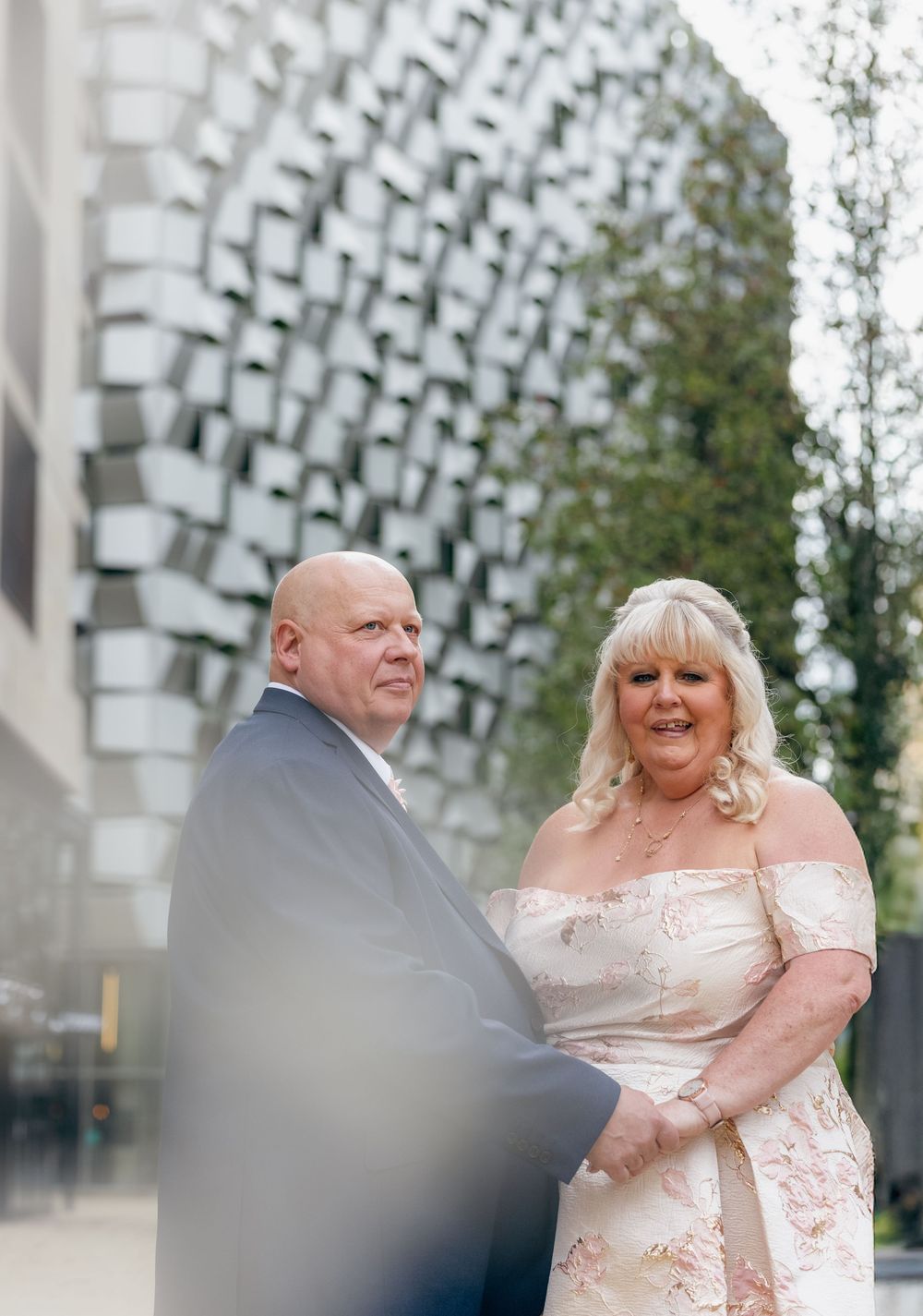 A couple poses together in formal attire against a modern architectural backdrop.
