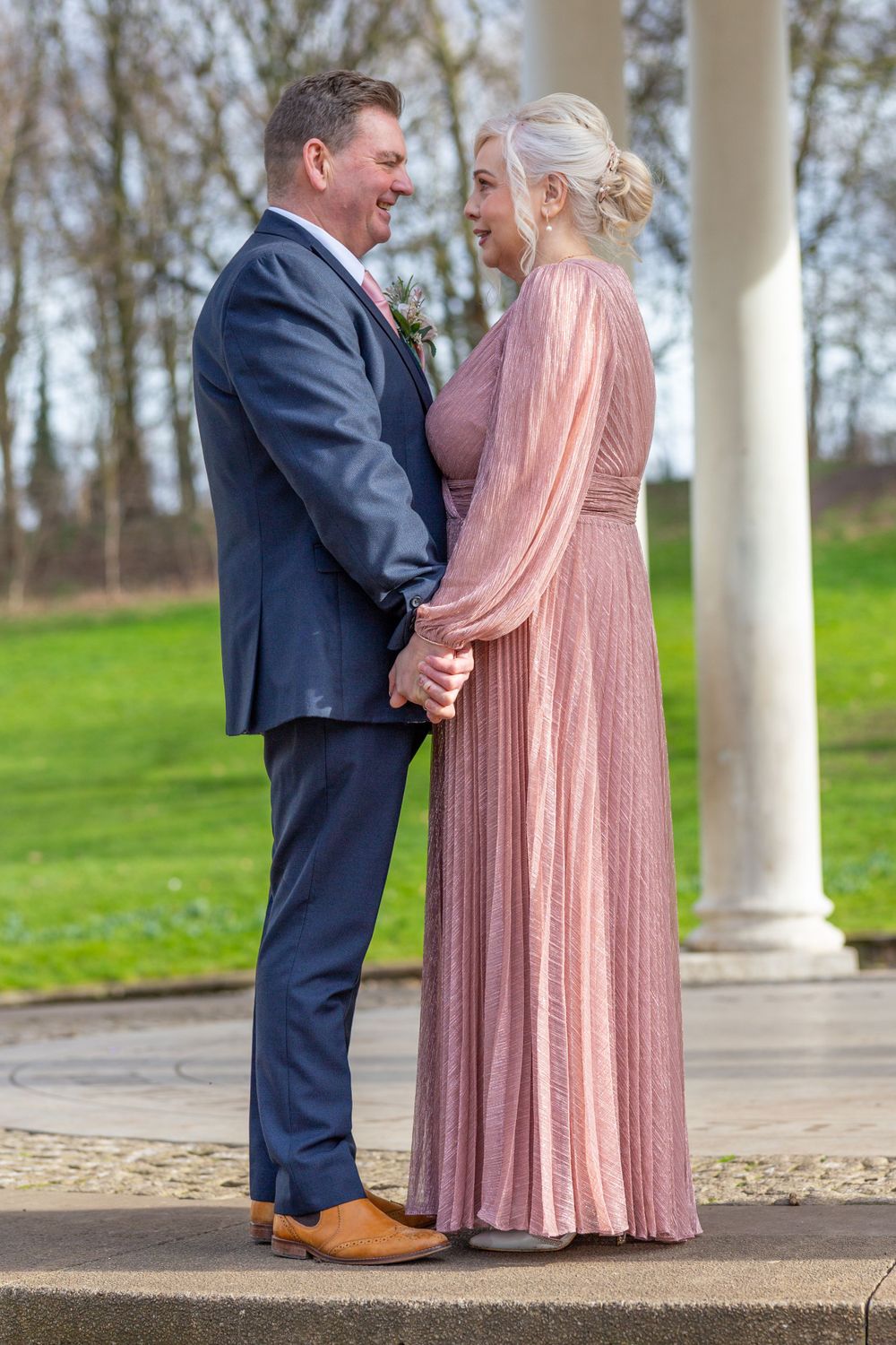 Romantic couple in navy suit and pink dress posing under white pillars outdoors.