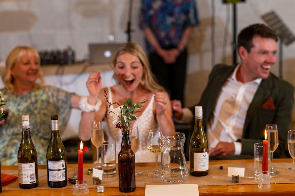 Wedding reception guests laugh and celebrate at a table filled with wine bottles and glasses.
