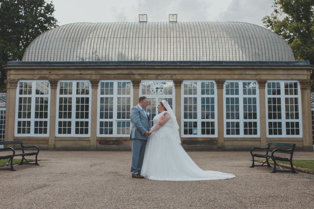 A couple poses in front of a curved glass conservatory building on their wedding day.