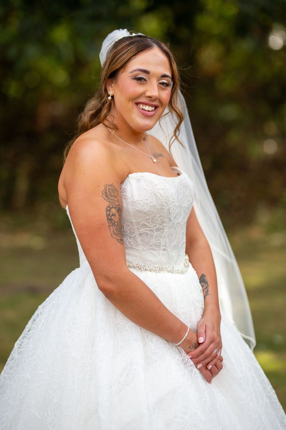 Smiling bride in strapless white lace ballgown with veil and floral tattoos poses outdoors.