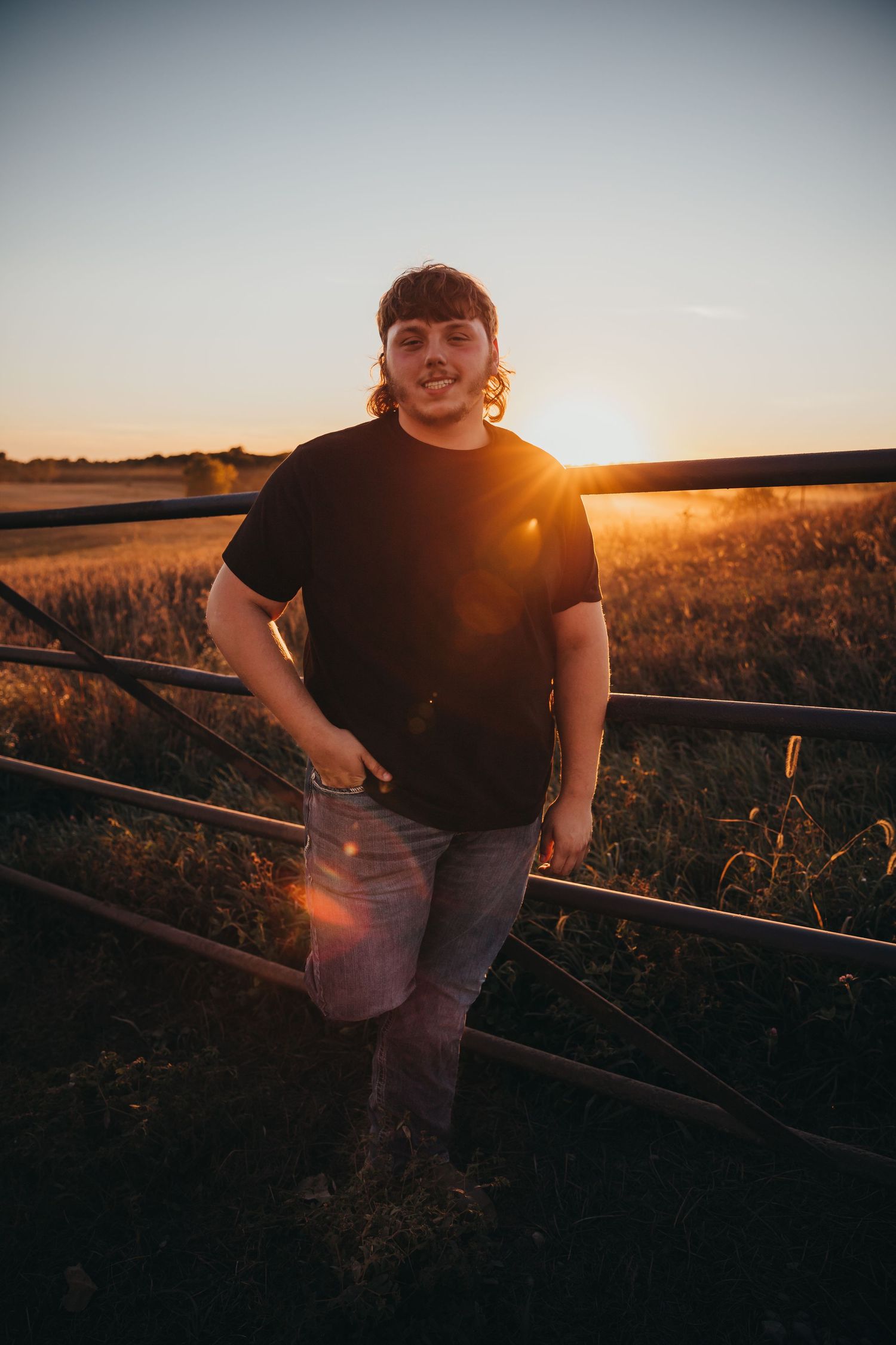Person in black t-shirt and jeans leans against metal fence at sunset in rural field with golden sunlight.
