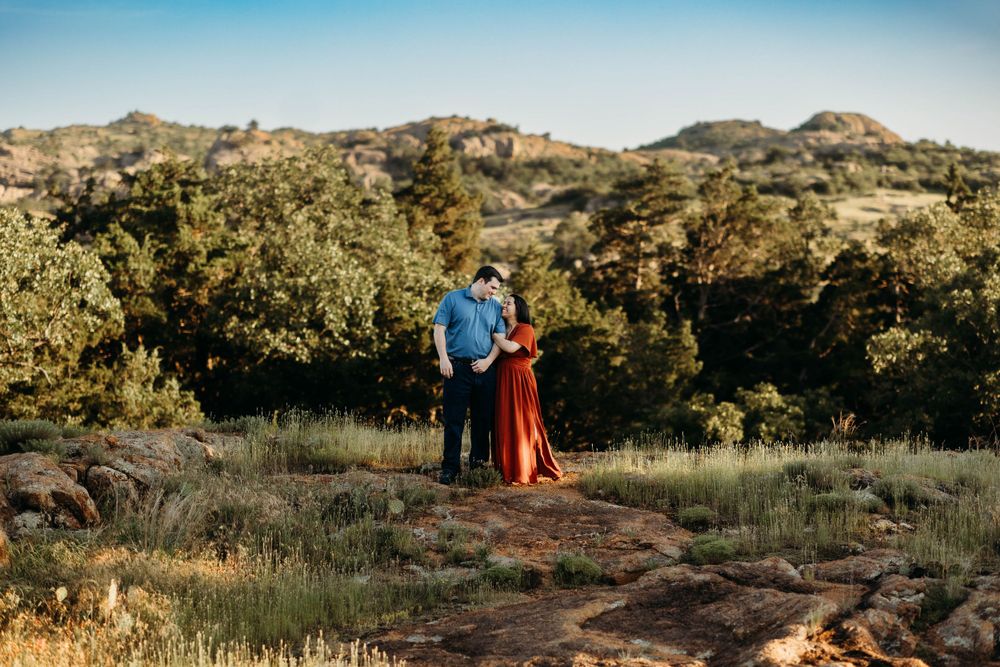 A couple stands on a hilltop at golden hour with mountains and green foliage in the background.