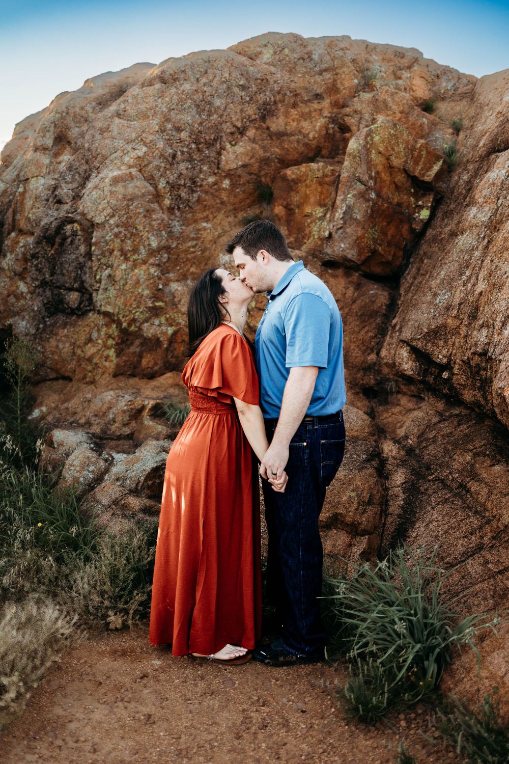 A couple shares a romantic moment on a dirt path against large rusty-colored rock formations under a bright blue sky.