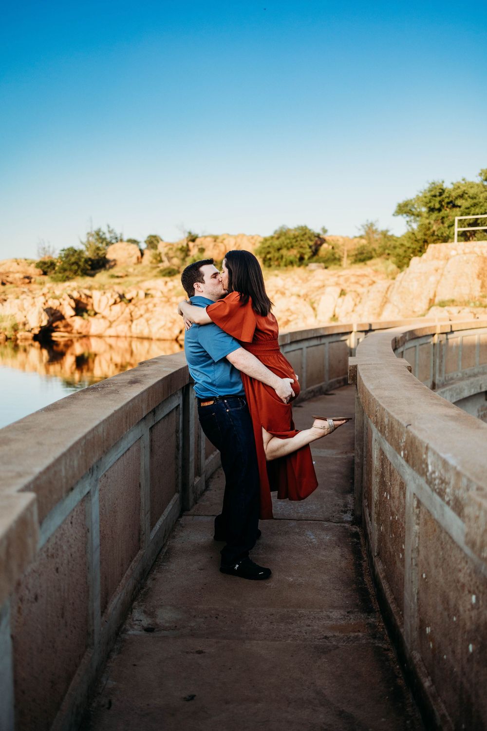 A romantic couple embraces on a concrete walkway overlooking a tranquil lake during golden hour.