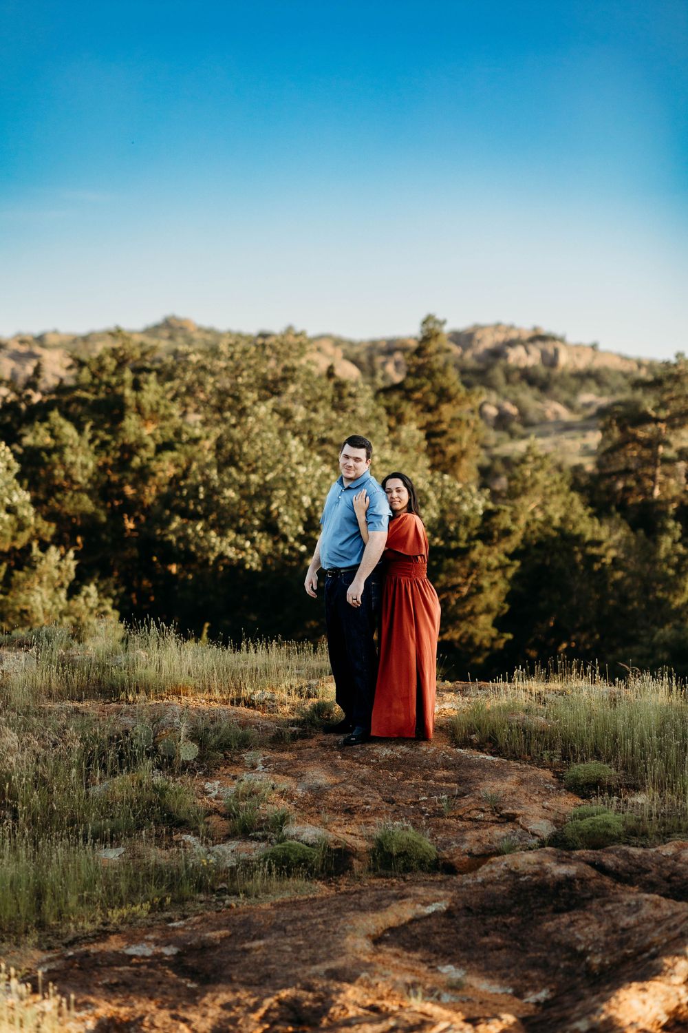 A couple standing together on a scenic hilltop trail surrounded by mountains and trees during golden hour lighting.