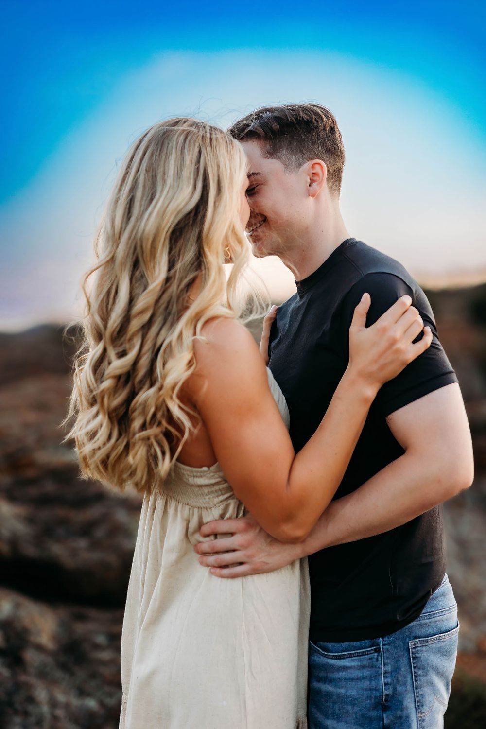 Romantic couple embracing outdoors against blue sky at sunset, with her blonde wavy hair and cream dress creating soft contrast.