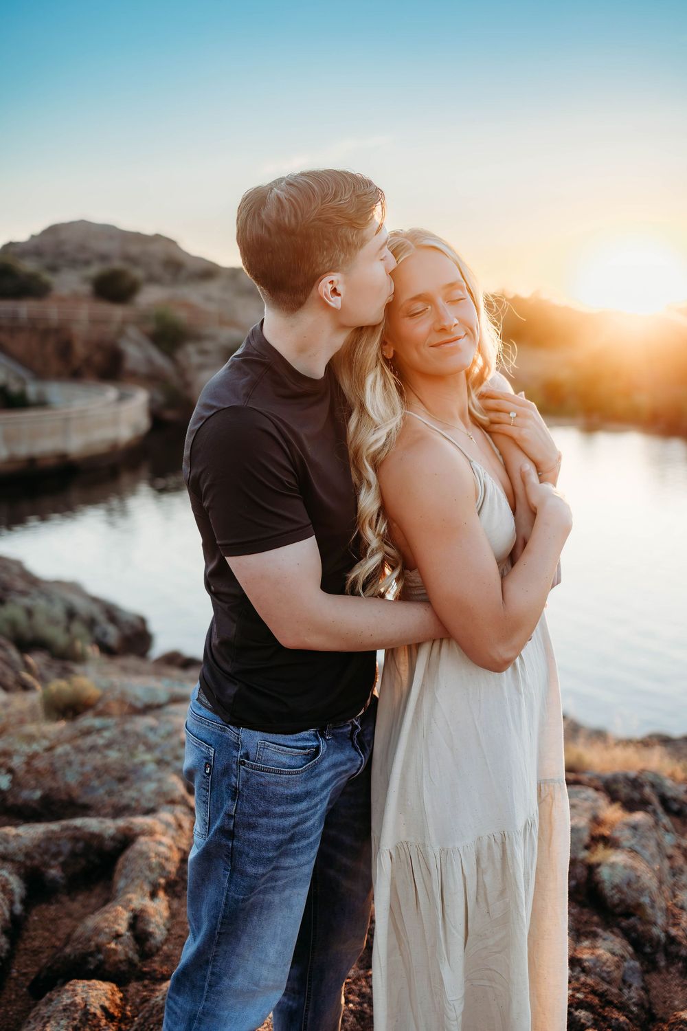 Romantic couple embraces at sunset by a rocky lake shore as golden light illuminates their tender moment.