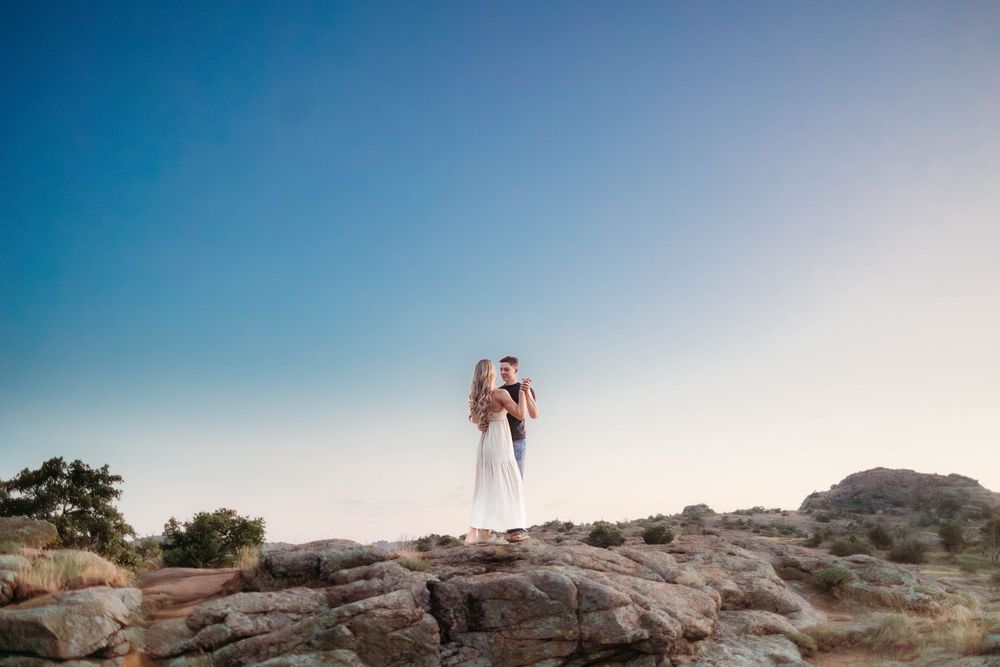 Two figures stand together on a rocky outcrop against a bright blue sky during golden hour.
