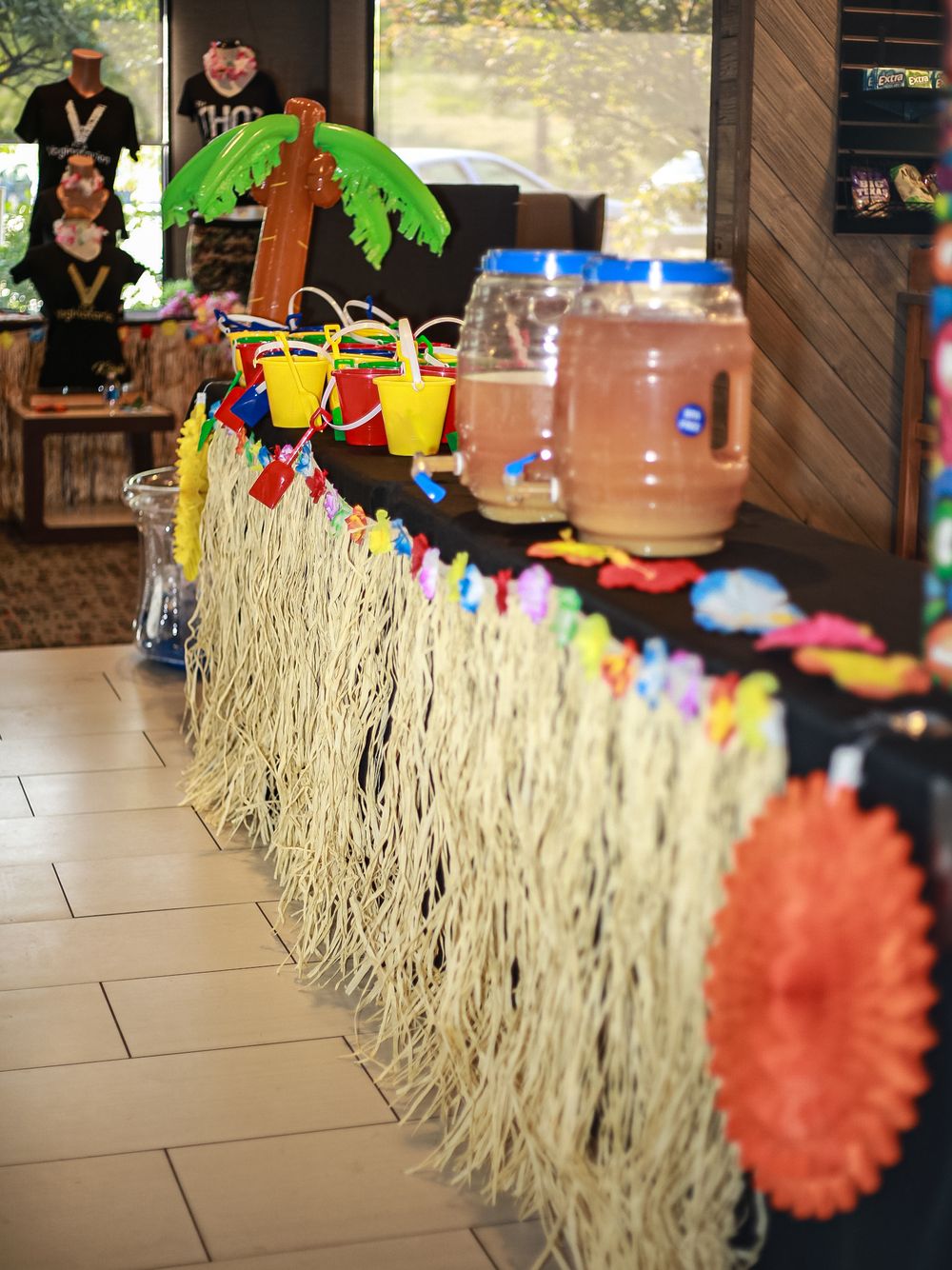A tropical-themed party bar setup with grass skirt decorations, colorful drinks, and a palm tree centerpiece.