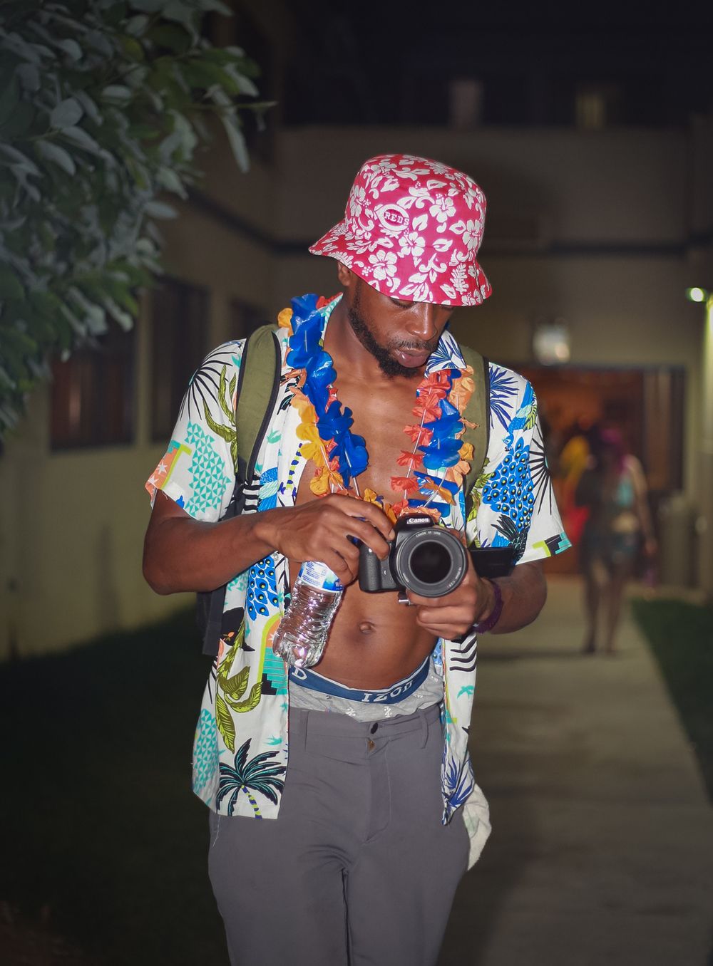 Photographer in tropical shirt and patterned bucket hat holds camera in dimly lit hallway with festive blue lei.