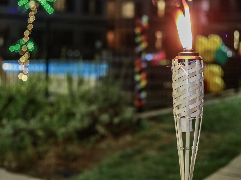 Tiki torch burning brightly at dusk with soft bokeh lights in the background and foliage visible.