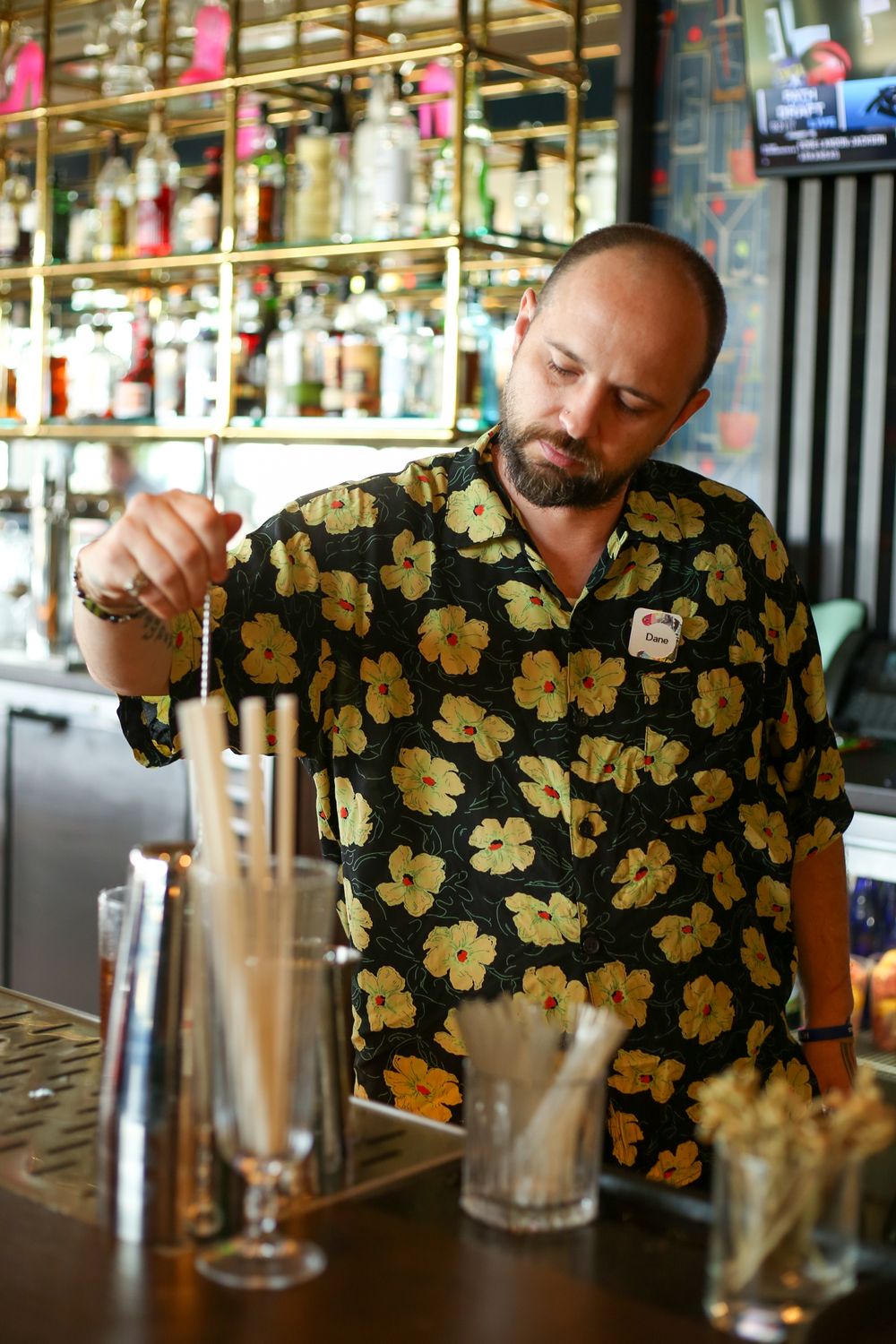 A bartender in a black and gold floral hawaiian shirt carefully measures ingredients at a well-stocked bar.