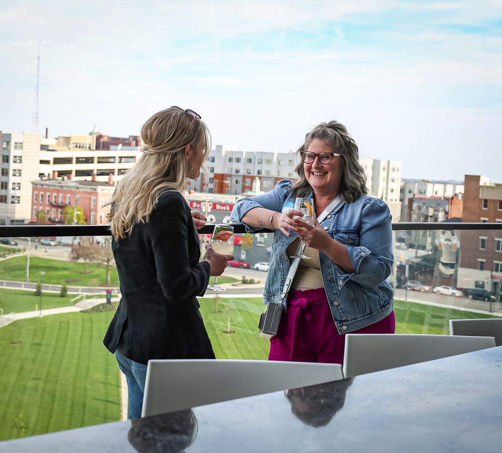 Two people share wine on a rooftop patio overlooking a sports field and urban buildings on a sunny day.