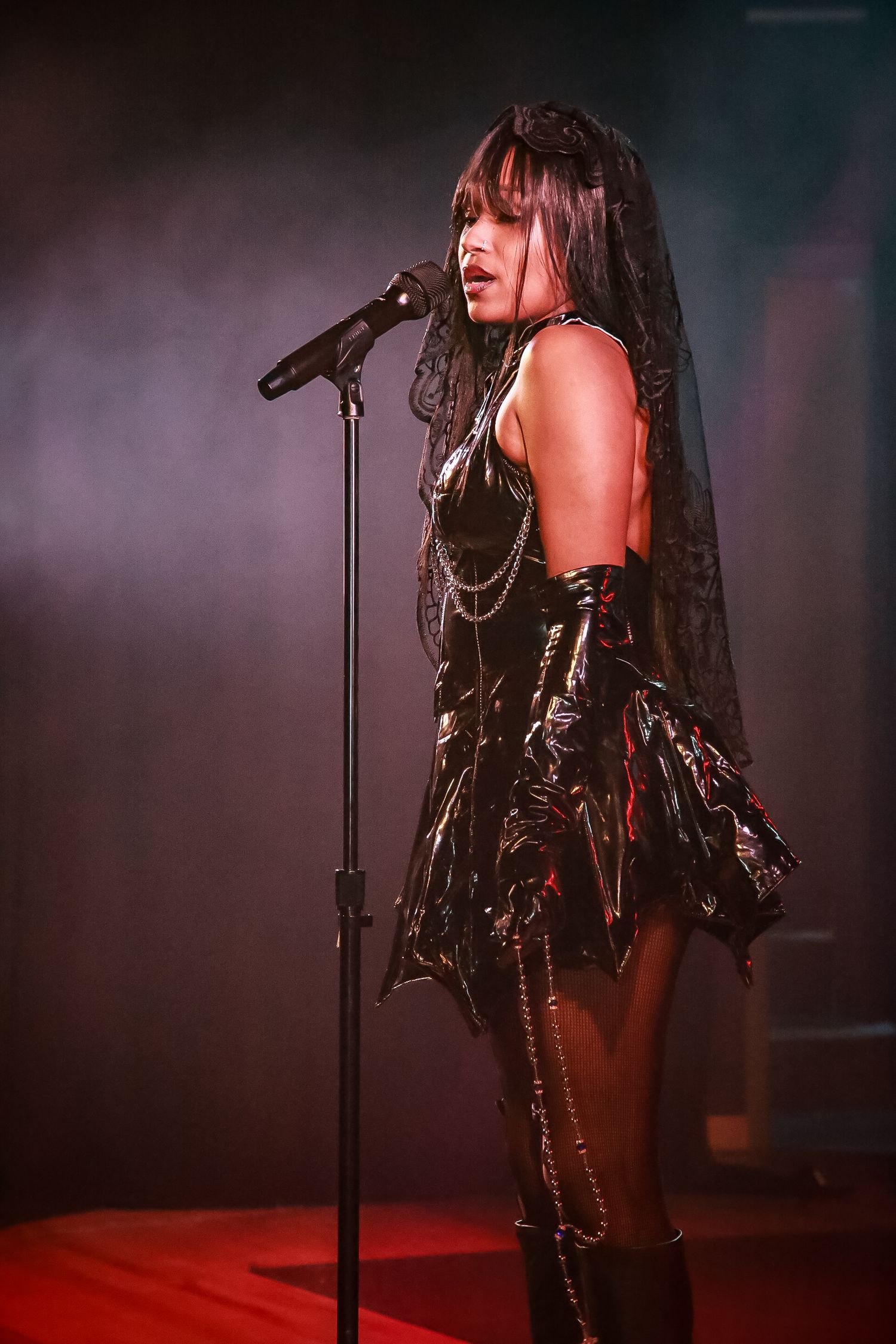 Performer in gothic black outfit with chains and veil sings into microphone on misty stage with dramatic red lighting.