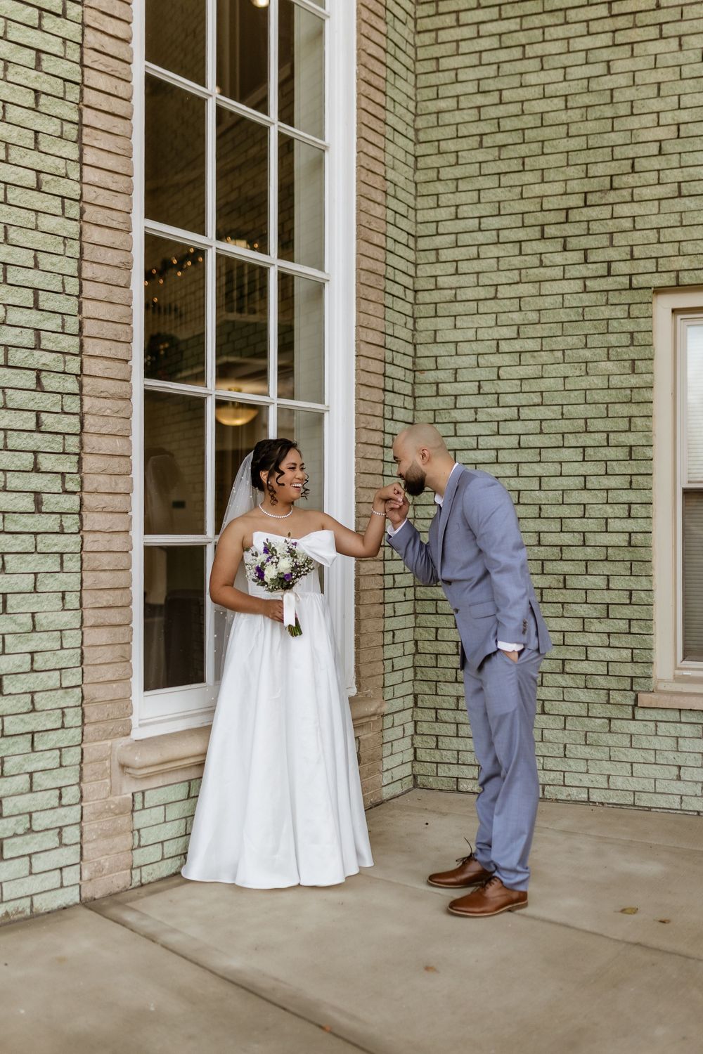 A romantic wedding moment against sage green brick walls as a couple shares a sweet courtship gesture.