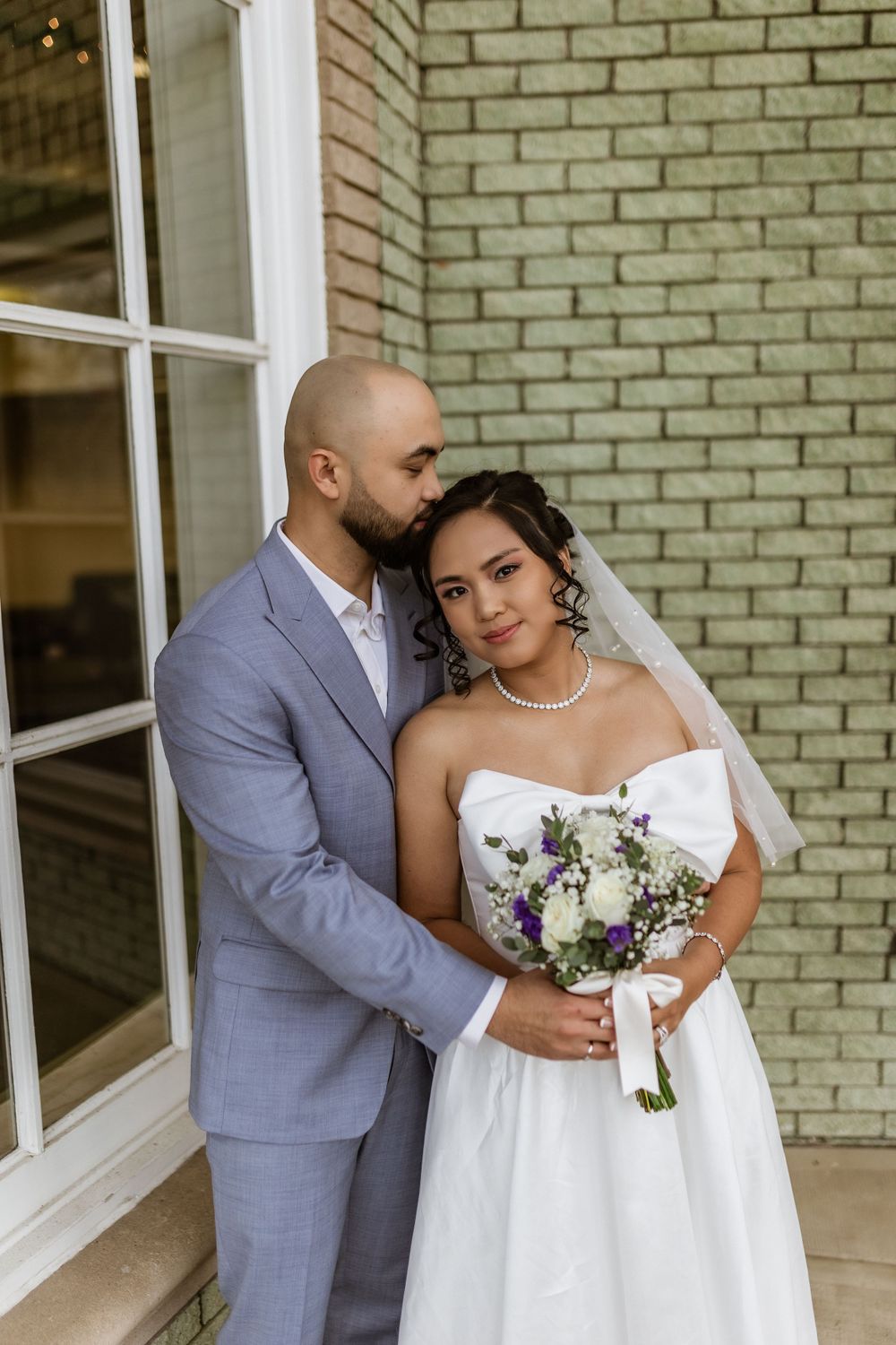 Newlyweds share an intimate moment in front of a mint green brick wall with classic white-framed windows.