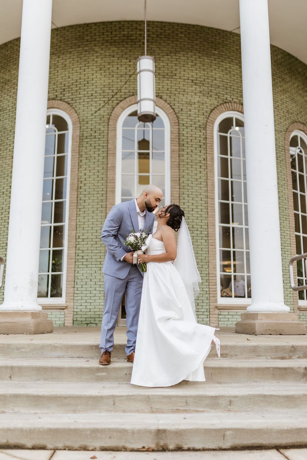 Wedding couple embraces on grand steps of a historic building with white columns and arched windows.