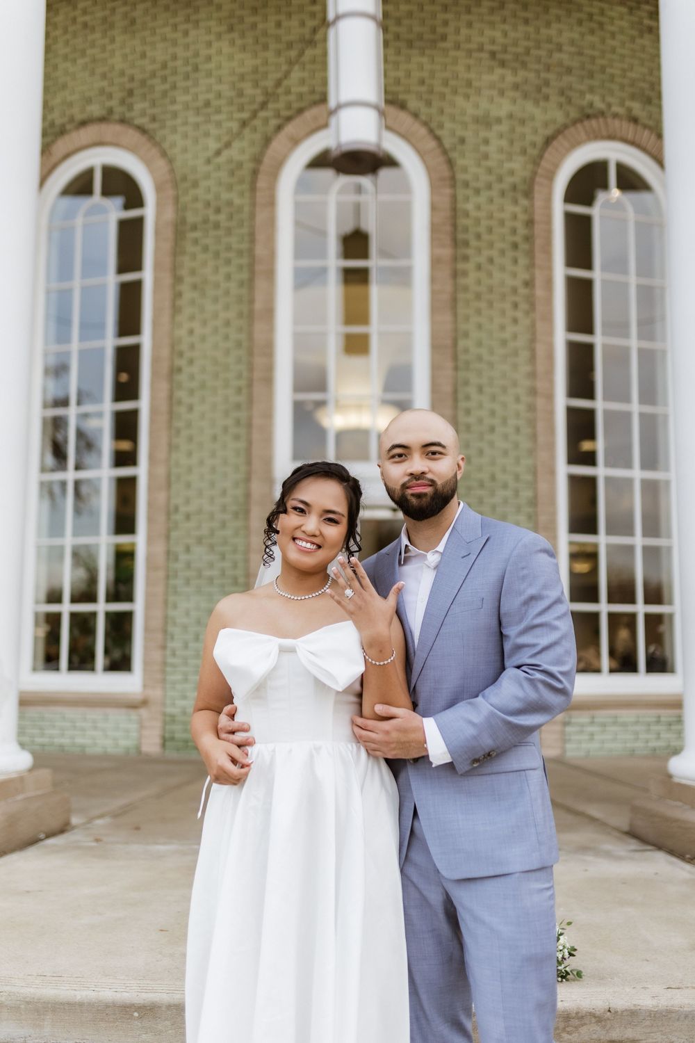 Happy newlyweds pose together in front of a charming brick building with tall arched windows.