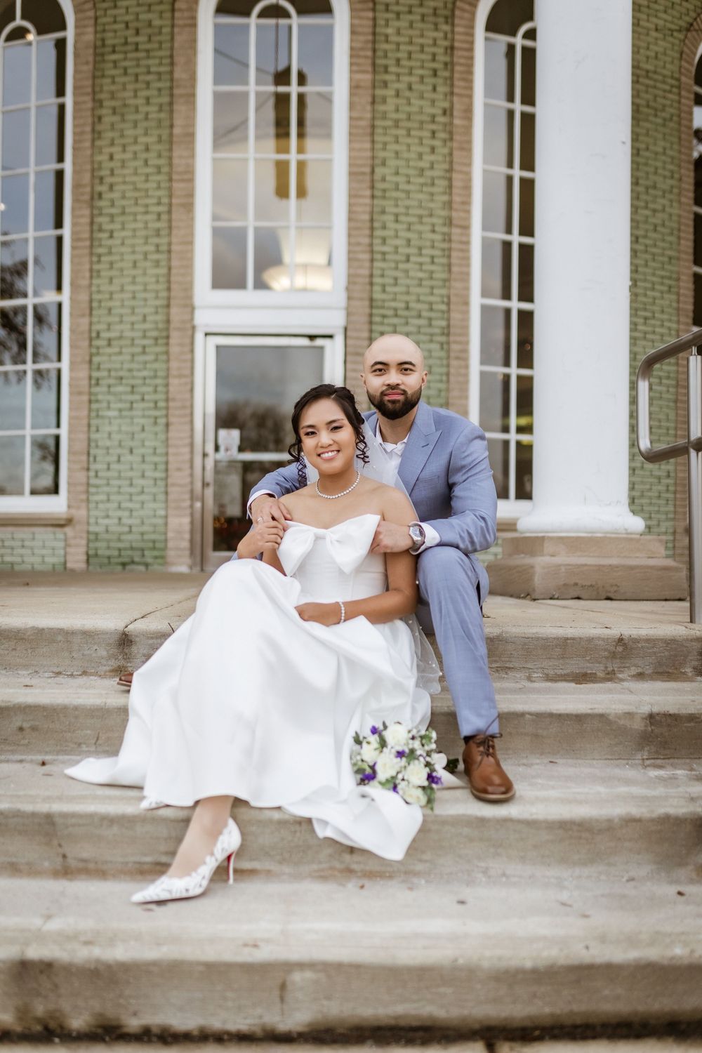 Couple shares a relaxed moment sitting on stone steps of a classic building with white columns.