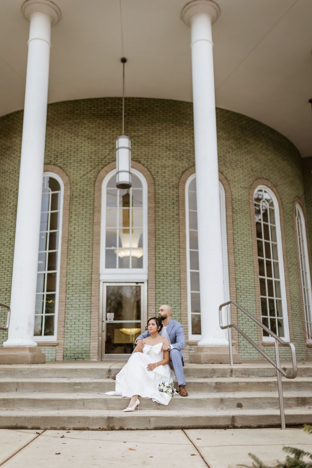 Wedding portrait on elegant stone steps with white columns and sage green brick architecture.