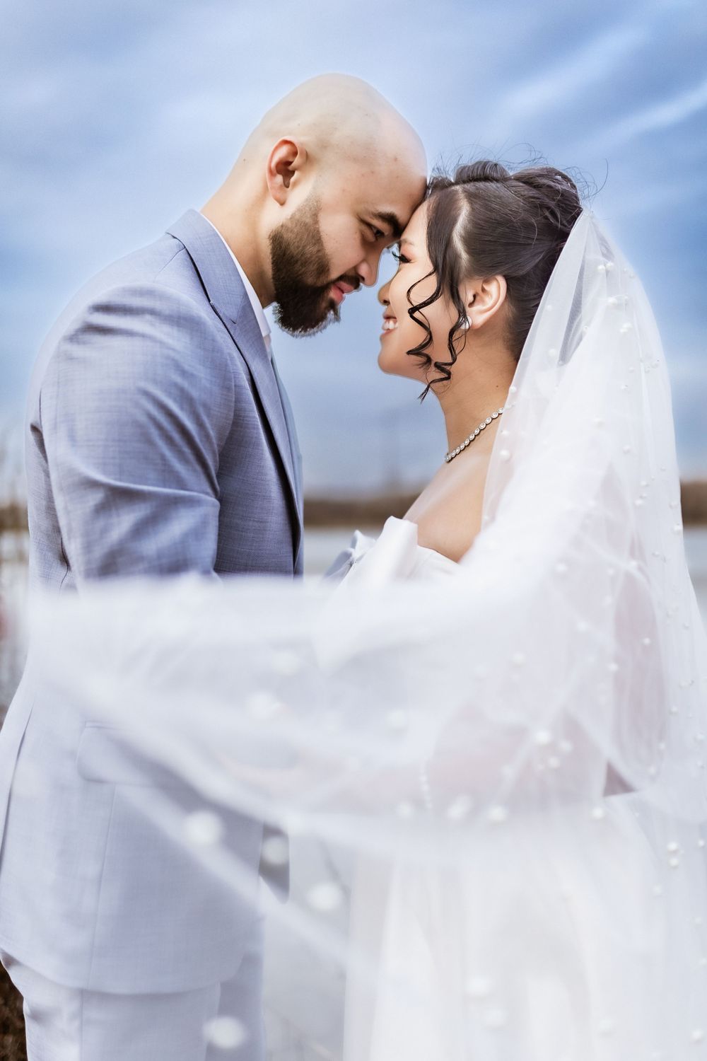 Romantic wedding portrait with flowing veil against soft blue sky.