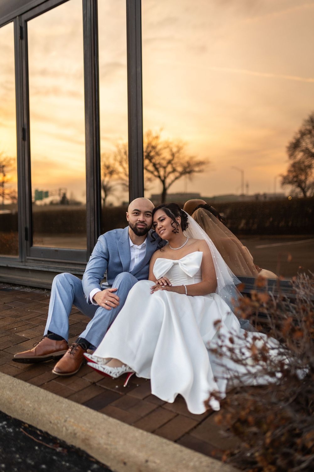 Wedding couple in light blue suit and white dress sitting together at sunset by glass building.