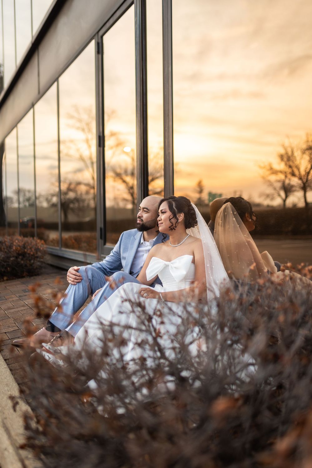 Newlyweds relaxing by modern glass building during golden hour sunset.