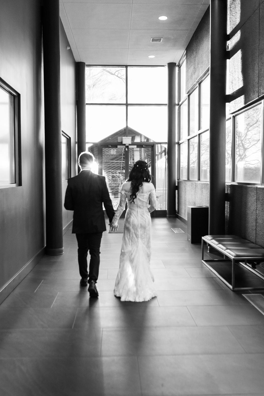 Bride and groom walking down modern hallway towards sunlit exit in black and white.
