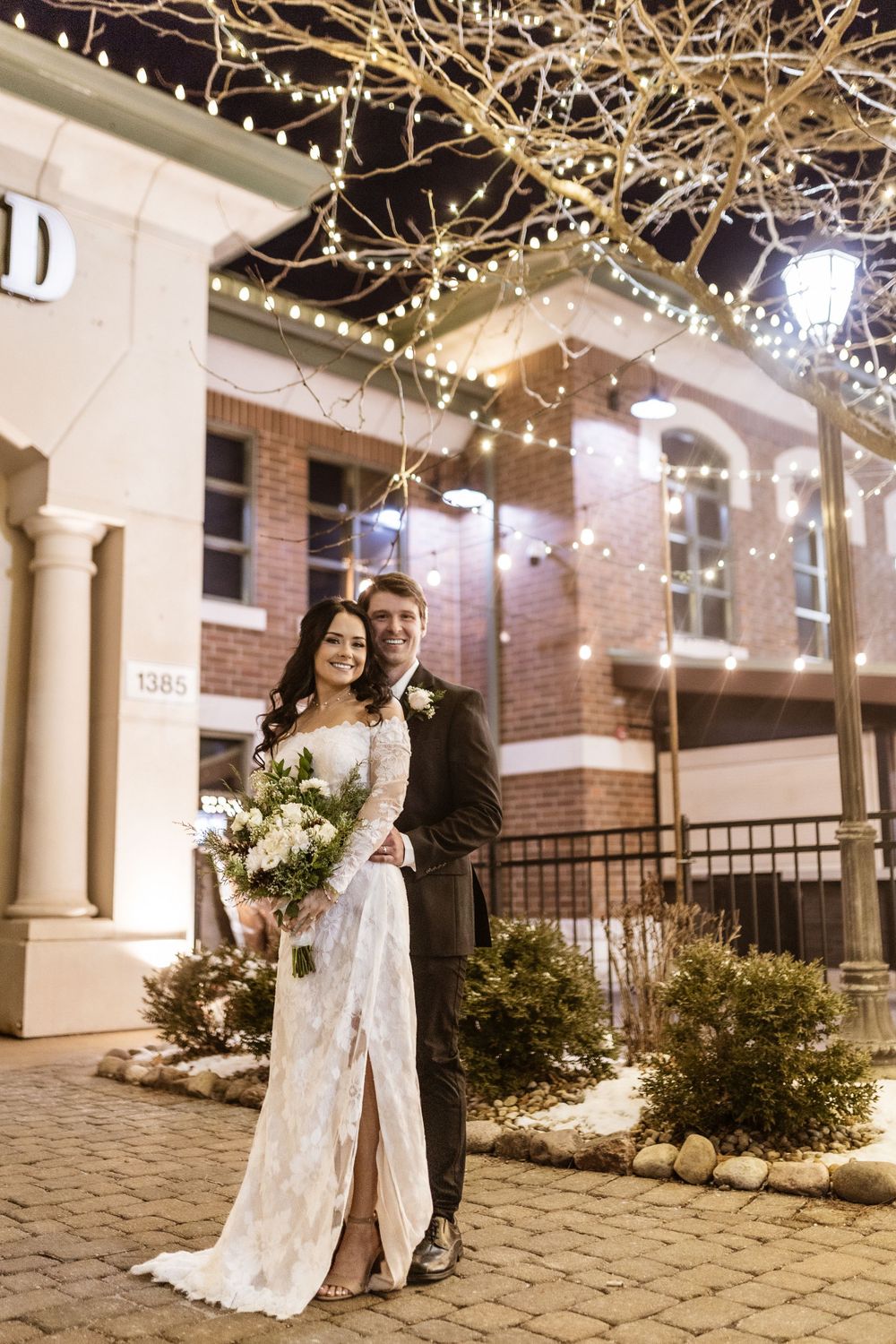 Elegant couple posing outside brick venue with string lights and columns.