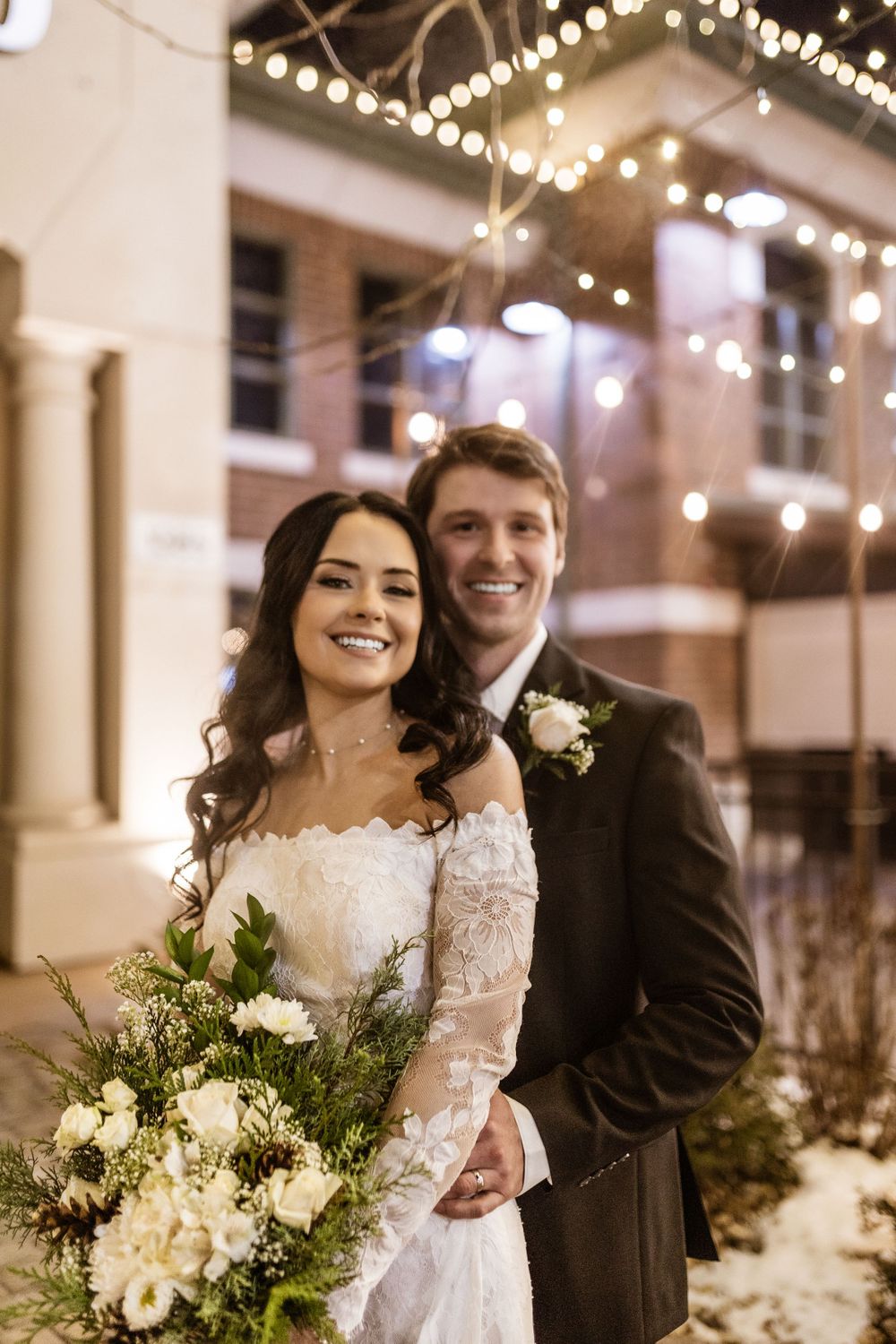 Happy bride holding white bouquet while sharing joyful moment with groom under string lights.