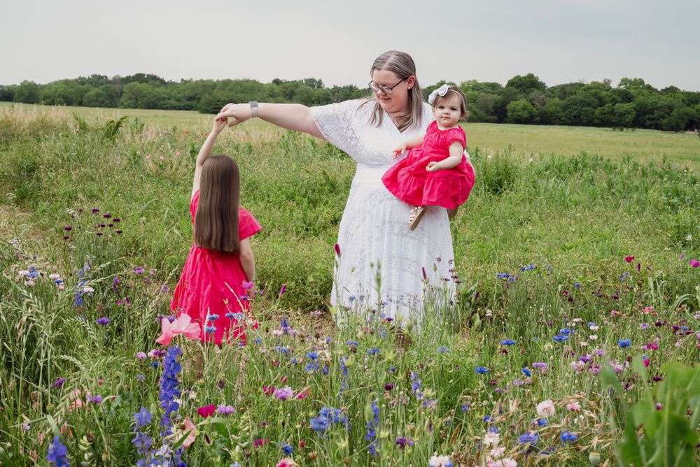 A woman in white holds two children wearing bright red dresses in a wildflower field.