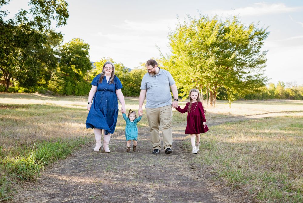 A family walks together on a dirt path surrounded by green trees on a sunny day.