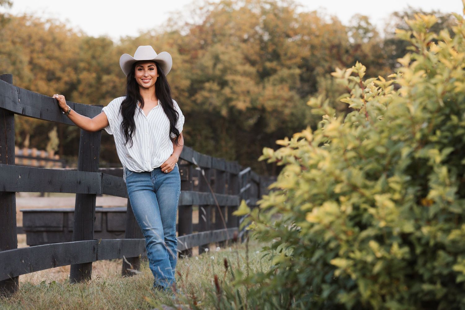 Casual country style with white hat and jeans leaning on wooden fence at sunset in rural setting.