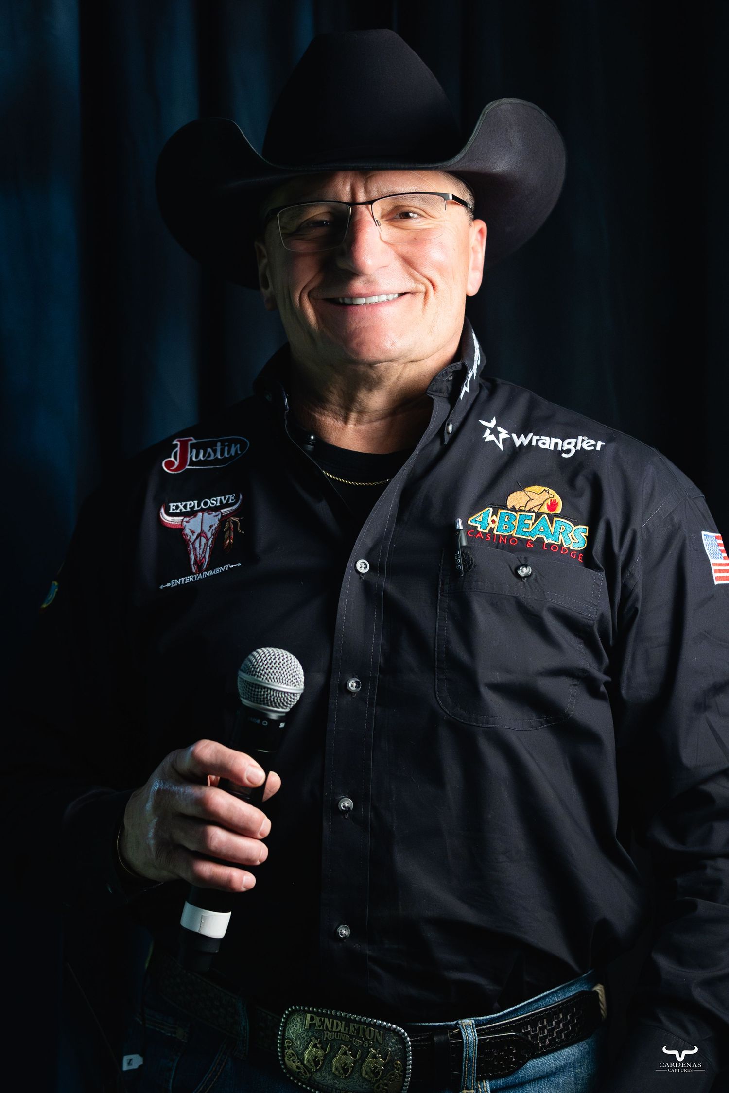 Rodeo announcer in black western jacket holding microphone with sponsor logos.