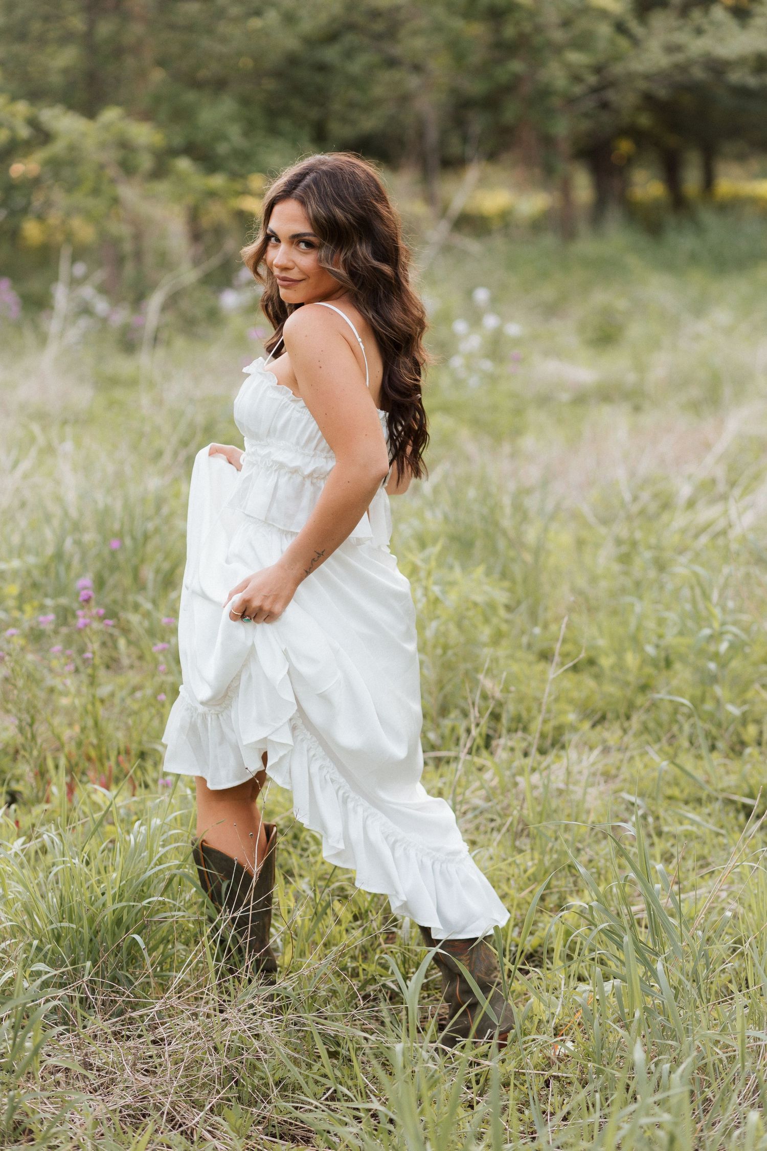 Woman in white ruffled dress and brown cowboy boots standing in a lush green meadow, smiling over her shoulder.