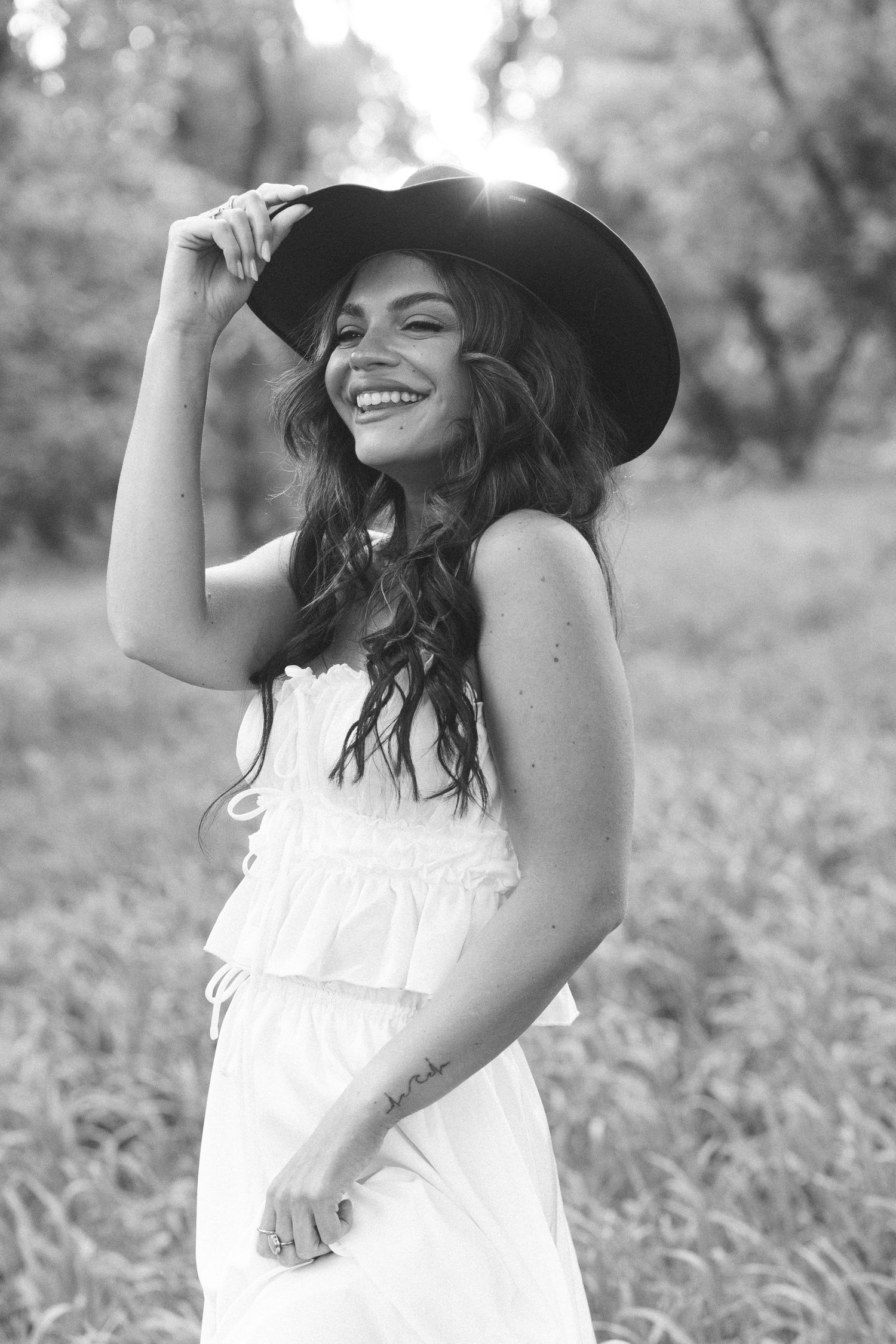 Black and white photo of a smiling woman in a white dress and wide-brim hat standing outdoors in a field.