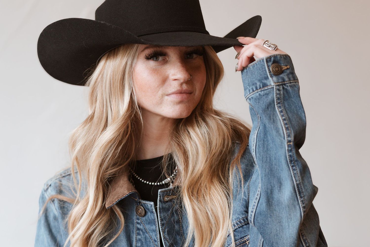 Person in western style wearing denim jacket and black cowboy hat with wavy blonde hair poses against neutral background.
